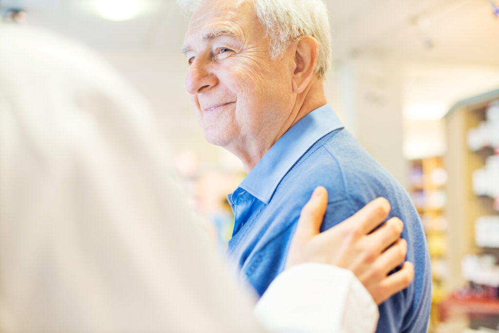 Arzt legt Hand auf die Schulter eines Patienten, der ältere Patient lächelt ihm zu