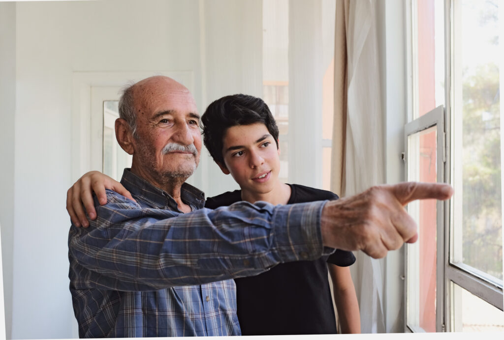 Alter bärtiger Mann steht zusammen mit seinem Enkel am Fenster und zeigt nach draussen